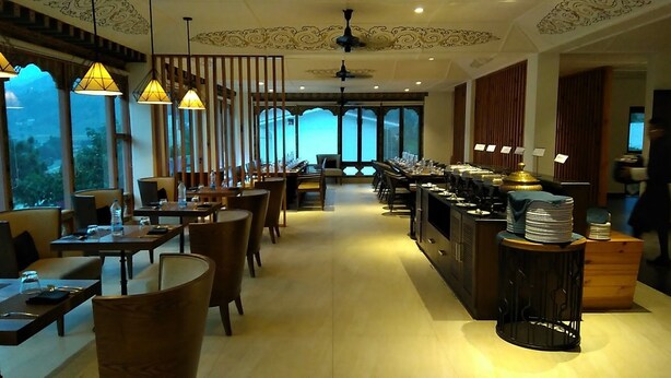 A restaurant with wooden tables and chairs, a counter with stacked plates, and large windows offering a view of the outdoors. - Khuru Resort (Punakha)