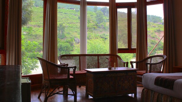 Une pièce dotée d’une grande fenêtre donnant sur un paysage verdoyant et luxuriant. On y trouve une table en bois, deux chaises et une boîte décorative.