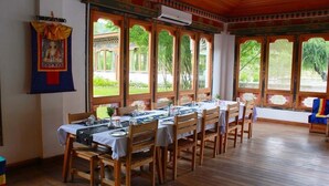 Une salle à manger avec une longue table dressée pour accueillir plusieurs personnes, de grandes fenêtres dotées de cadres en bois traditionnels et une tapisserie suspendue au mur.