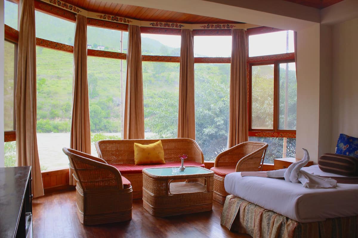 A room with wicker furniture, a bed, and large windows offering a view of greenery.