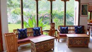 Une pièce meublée de façon traditionnelle avec des meubles en bois, une grande fenêtre donnant sur un paysage verdoyant et une petite table aux motifs complexes.