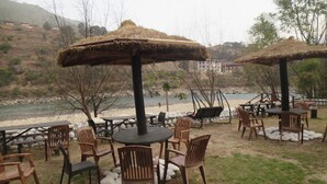 Un espace extérieur aménagé avec des tables et des chaises, des parasols en chaume et une vue sur une rivière et des collines en arrière-plan.