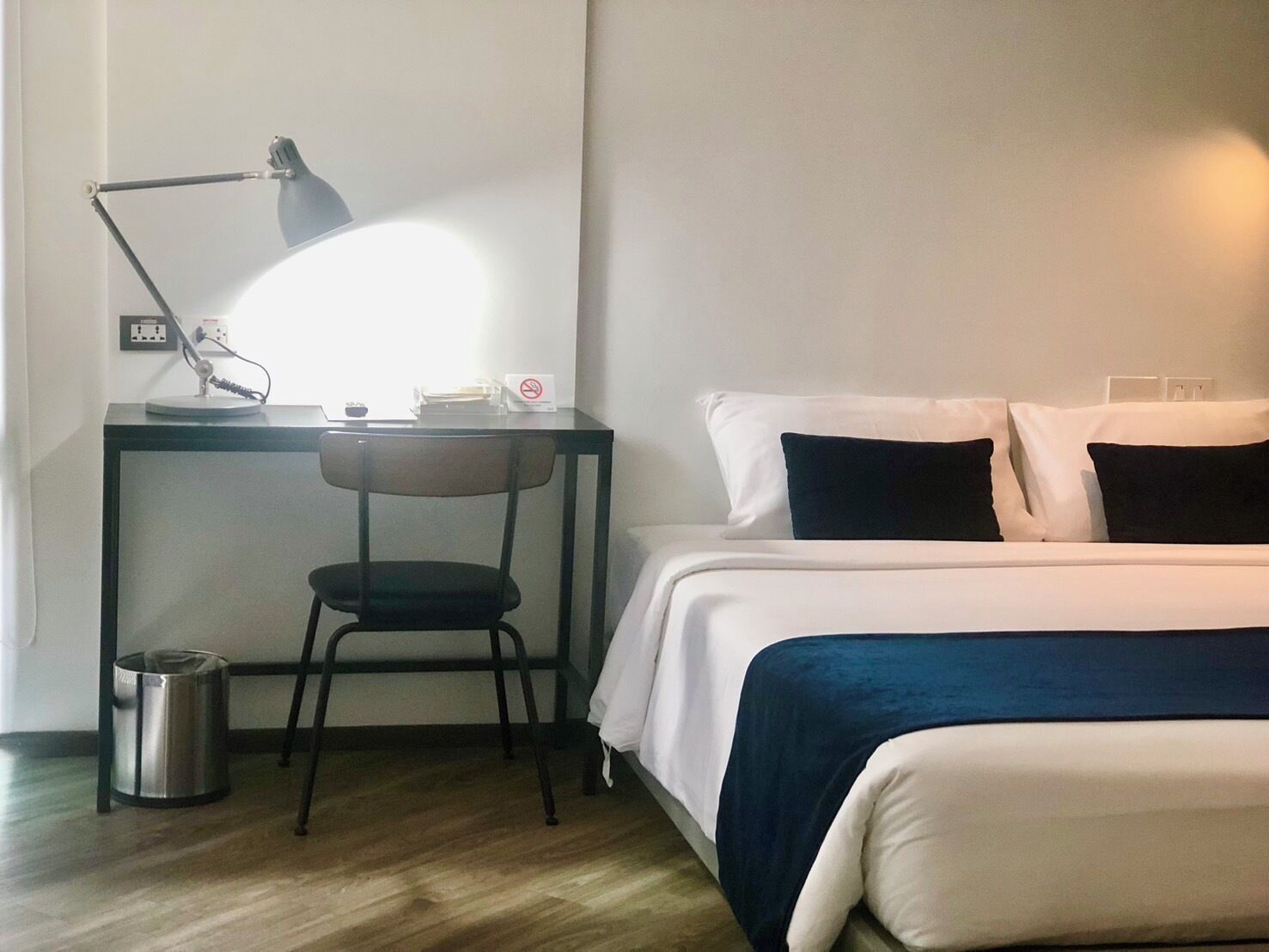 A hotel room with a bed, a desk with a lamp, a chair, and a trash bin.