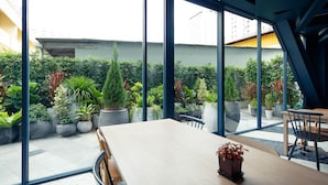 A modern indoor space with a large table and chairs, surrounded by large windows offering views of a rooftop garden with various plants and a building in the background. - Klub Hotel (Bangkok)
