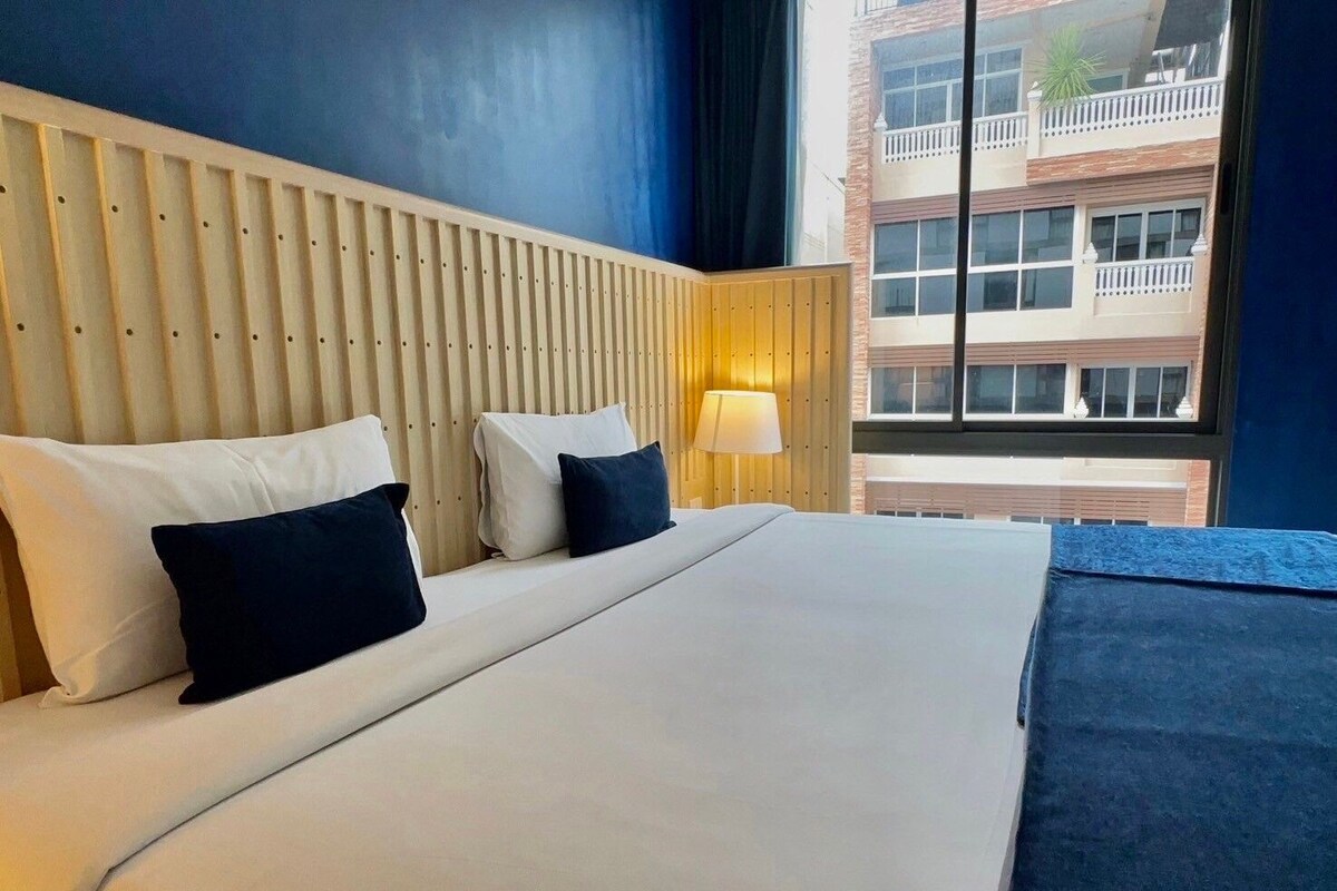 A hotel room with a bed, pillows, a nightstand, a lamp, and a view of a building through a window.