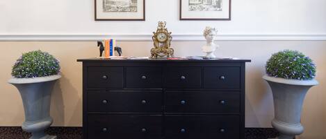 A black dresser with drawers, two framed black and white photographs on the wall, a gold clock, and two potted plants.