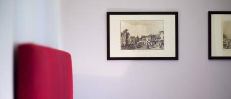 Una habitación con una silla roja, una obra de arte en blanco y negro de un paisaje urbano enmarcada, y una lámpara de mesa.