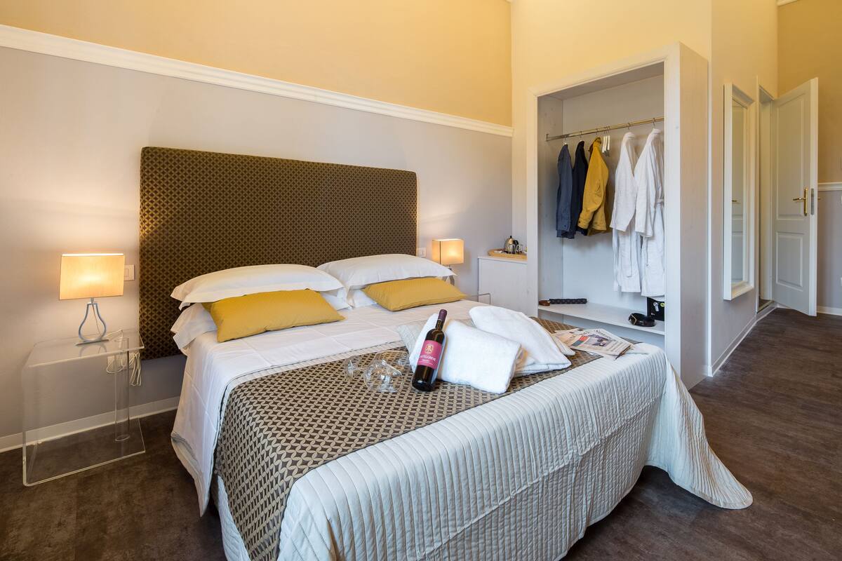 A hotel room with a large bed, a bedside table with a lamp, a robe rack with towels, and a door leading to another room.