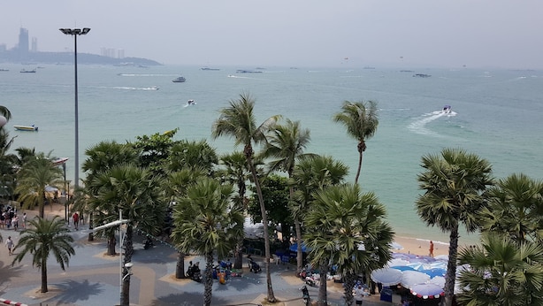 A beach with palm trees, a paved walkway, and boats in the water. - Charming Inn Hotel (Pattaya)