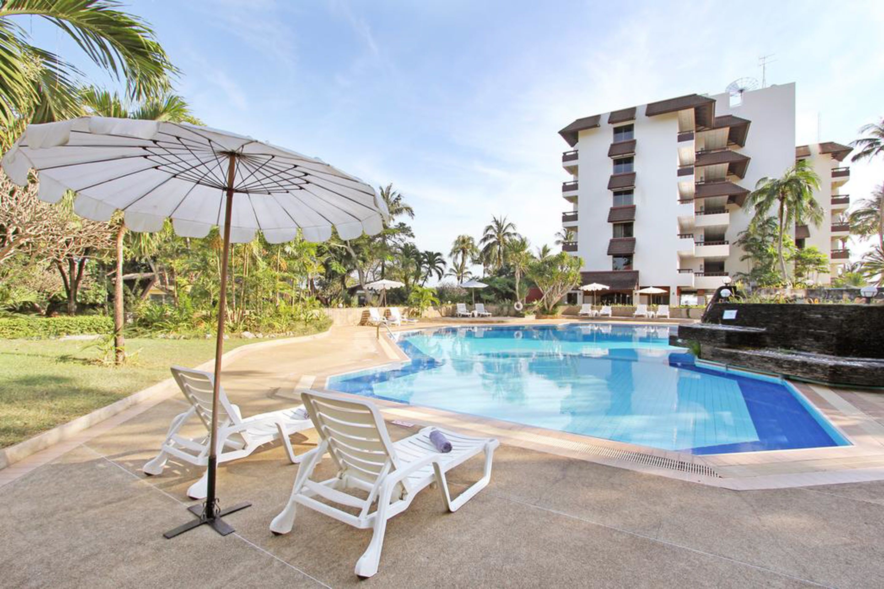 A swimming pool with lounge chairs and an umbrella, surrounded by palm trees and a multi-story building.