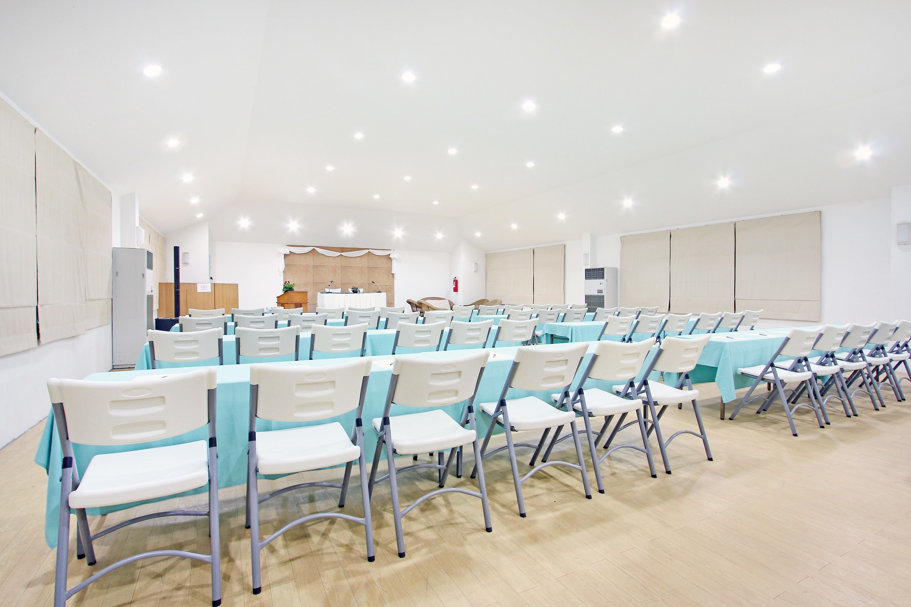 A conference room with rows of white chairs and tables, a stage with a podium, and a kitchen area in the background.