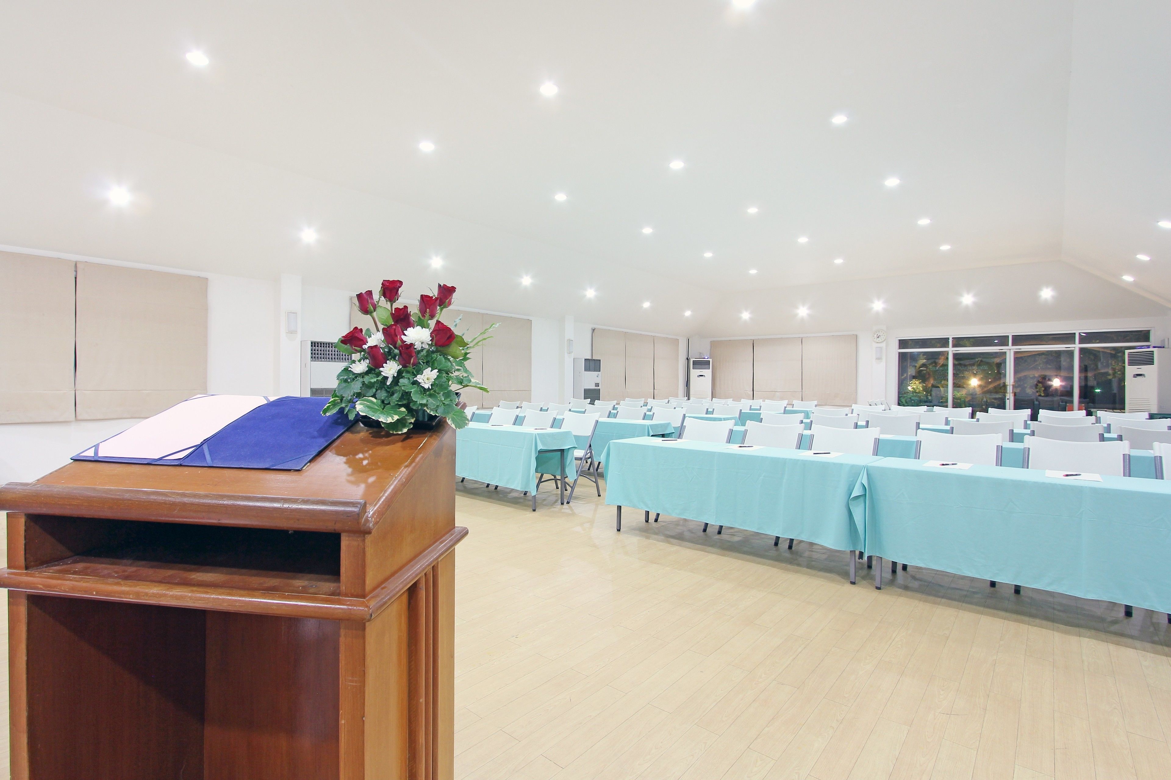 A conference room with rows of tables and chairs, a podium with a blue folder, and a floral arrangement.