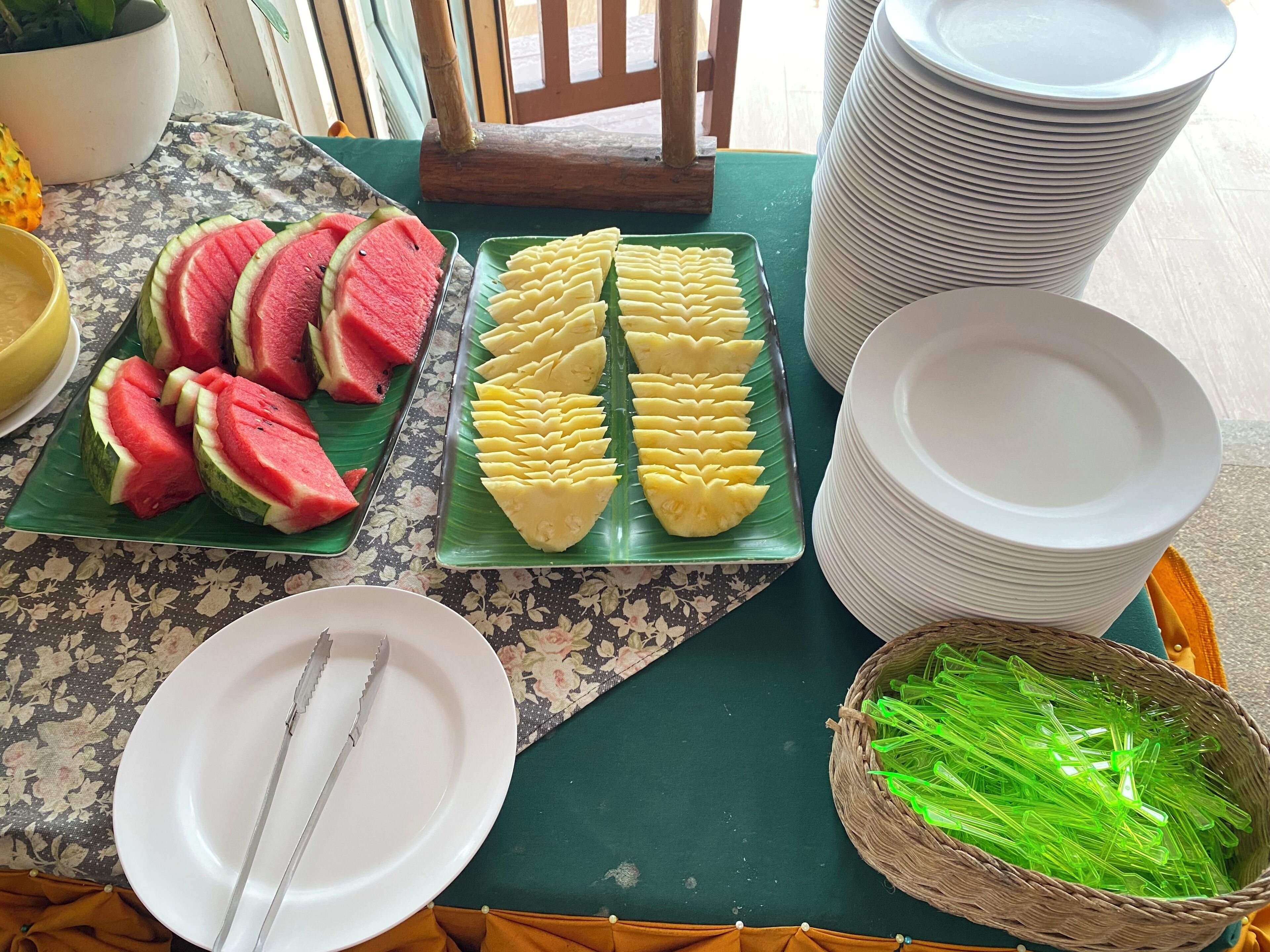 A table with plates of watermelon and pineapple slices, a stack of white plates, and a basket of green straws.