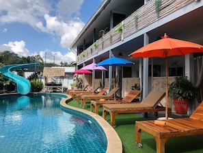 A swimming pool with lounge chairs and umbrellas, a slide, and a building with balconies. - Samui Zenity Hotel (Koh Samui)