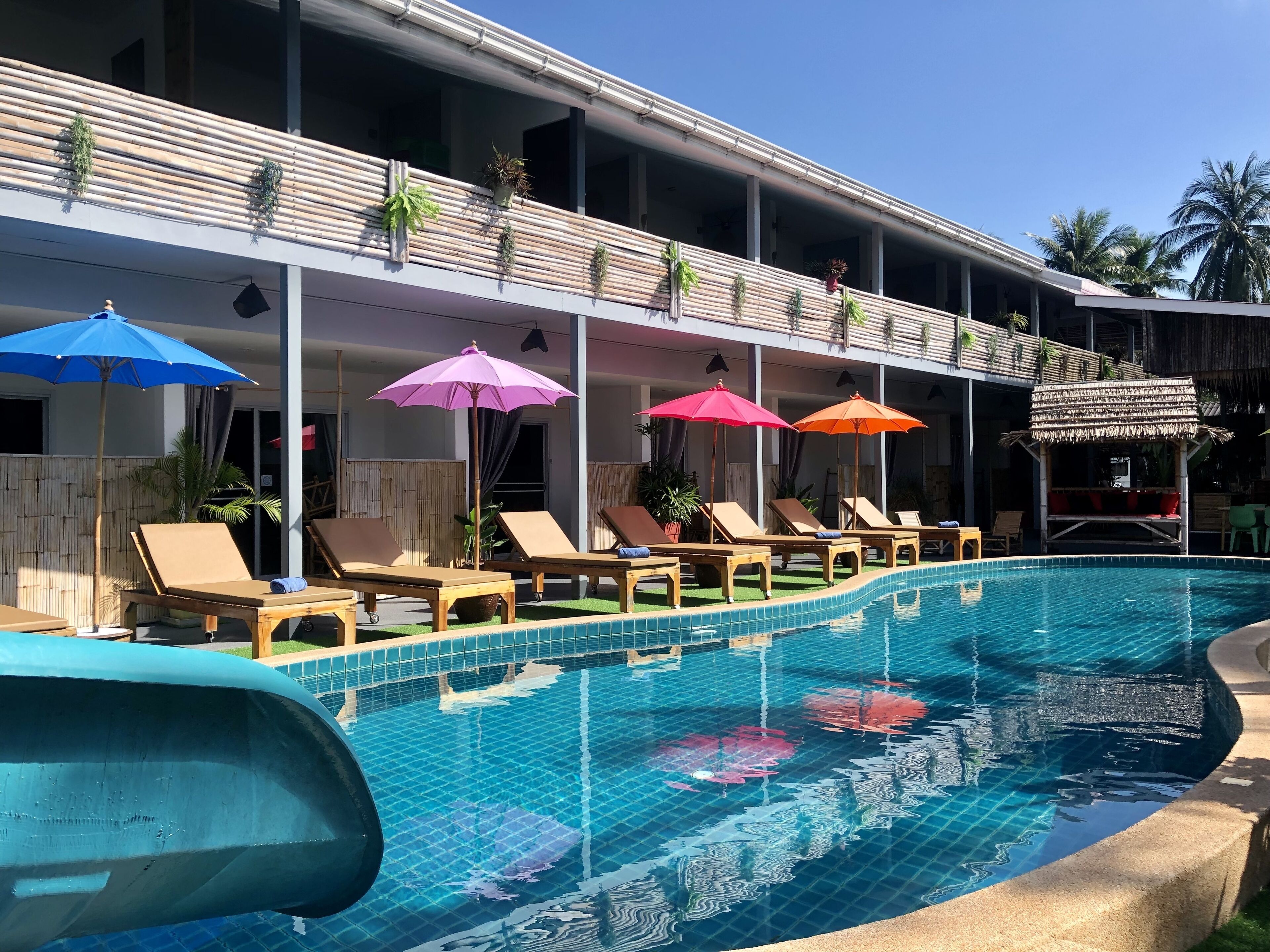 A pool area with lounge chairs and umbrellas, a covered seating area, and a building with a bamboo exterior.