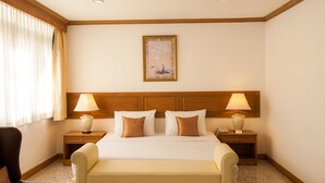 Une chambre d’hôtel avec un grand lit, deux tables de chevet avec des lampes, une tête de lit en bois, un tableau encadré au mur et une chaise.