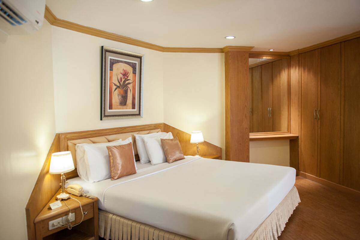 Une chambre d’hôtel avec un grand lit, une tête de lit en bois, deux tables de chevet avec des lampes, un tableau encadré au mur et des armoires en bois.