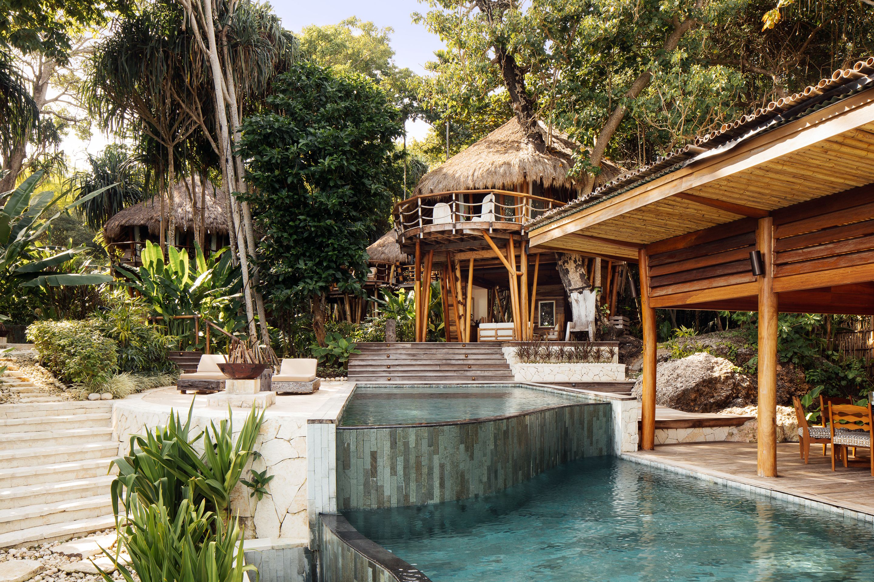 A pool area with a wooden deck, a thatched-roof structure, and lush greenery.