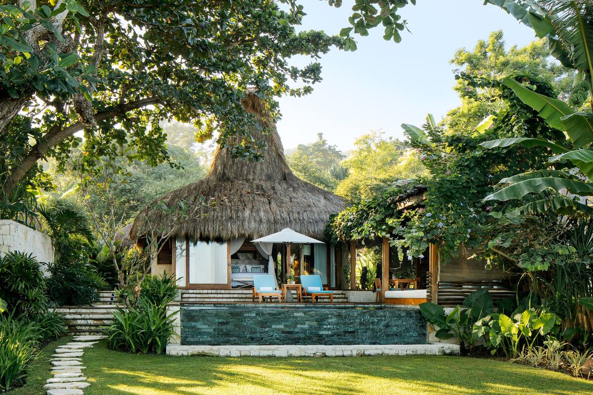 Un bungalow au toit de chaume, doté d’une piscine, entouré d’une végétation luxuriante et accessible par un sentier en pierre.