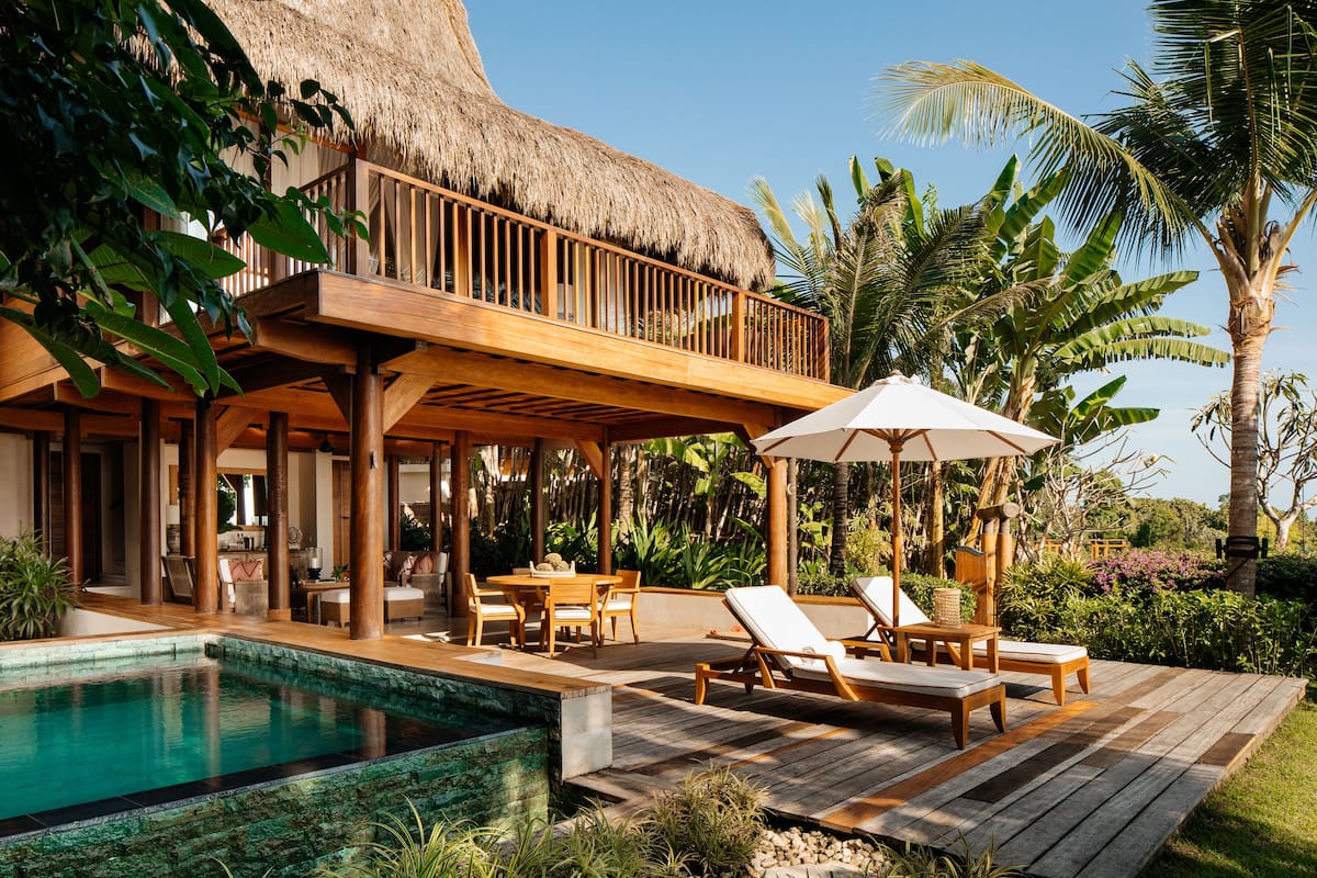 Une terrasse en bois avec des chaises longues et une piscine, entourée de plantes tropicales et d’une structure à toit de chaume.