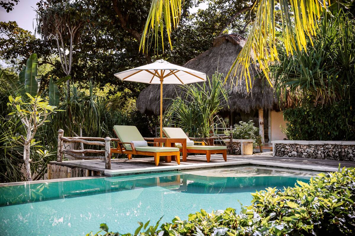 Un espace piscine doté de chaises longues et d’un parasol, entouré d’une végétation luxuriante et d’une structure à toit de chaume.