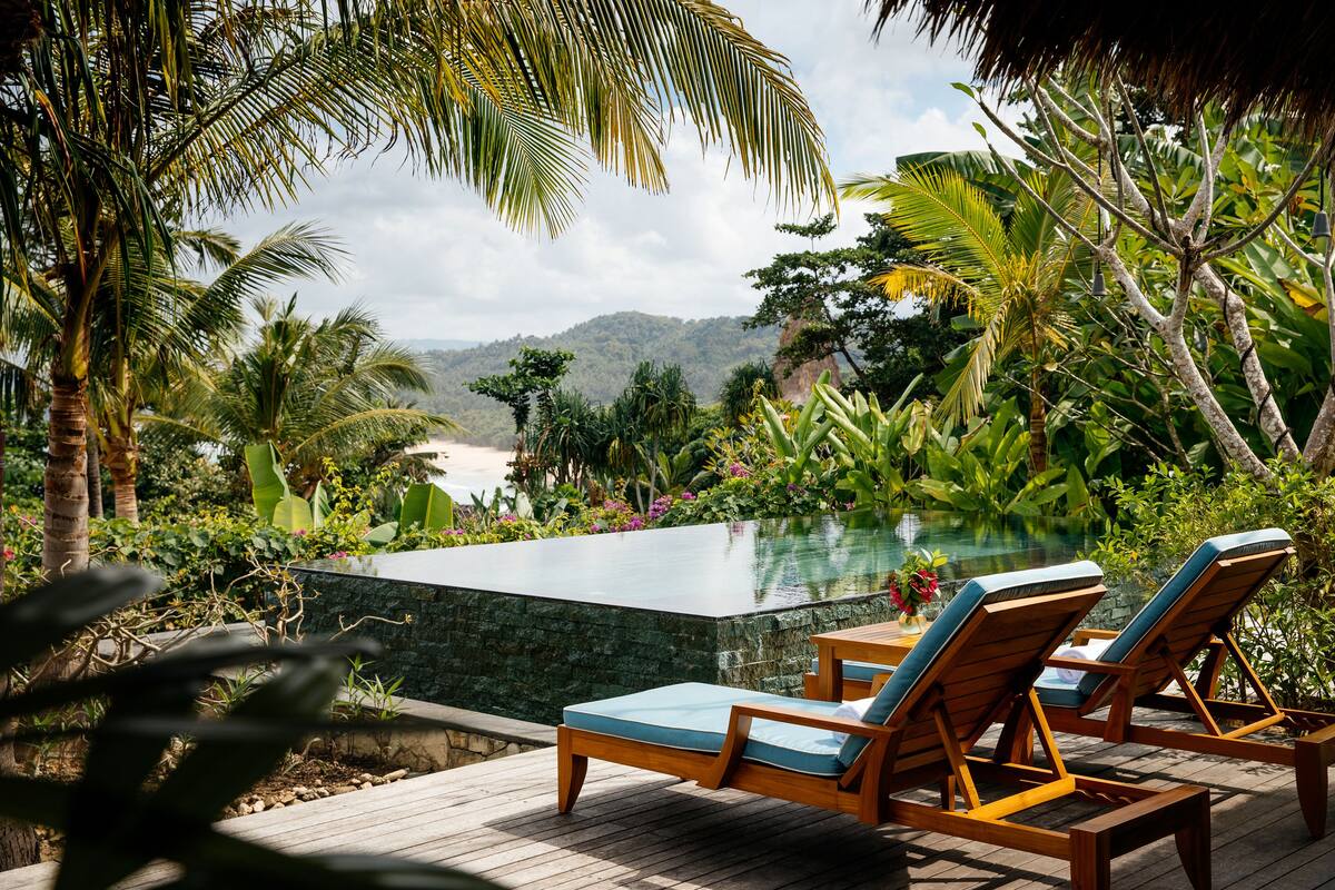 Un espace aménagé au bord de la piscine, avec des chaises longues en bois et une piscine en pierre, entouré de plantes tropicales et offrant une vue sur des collines lointaines.