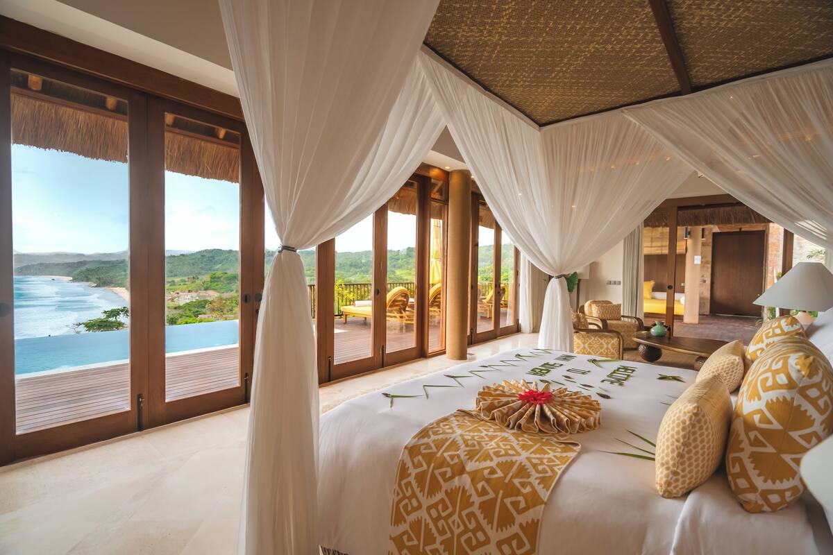 Une grande chambre avec vue sur l’océan, un balcon doté de mobilier d’extérieur et un grand lit agrémenté d’oreillers à motifs.