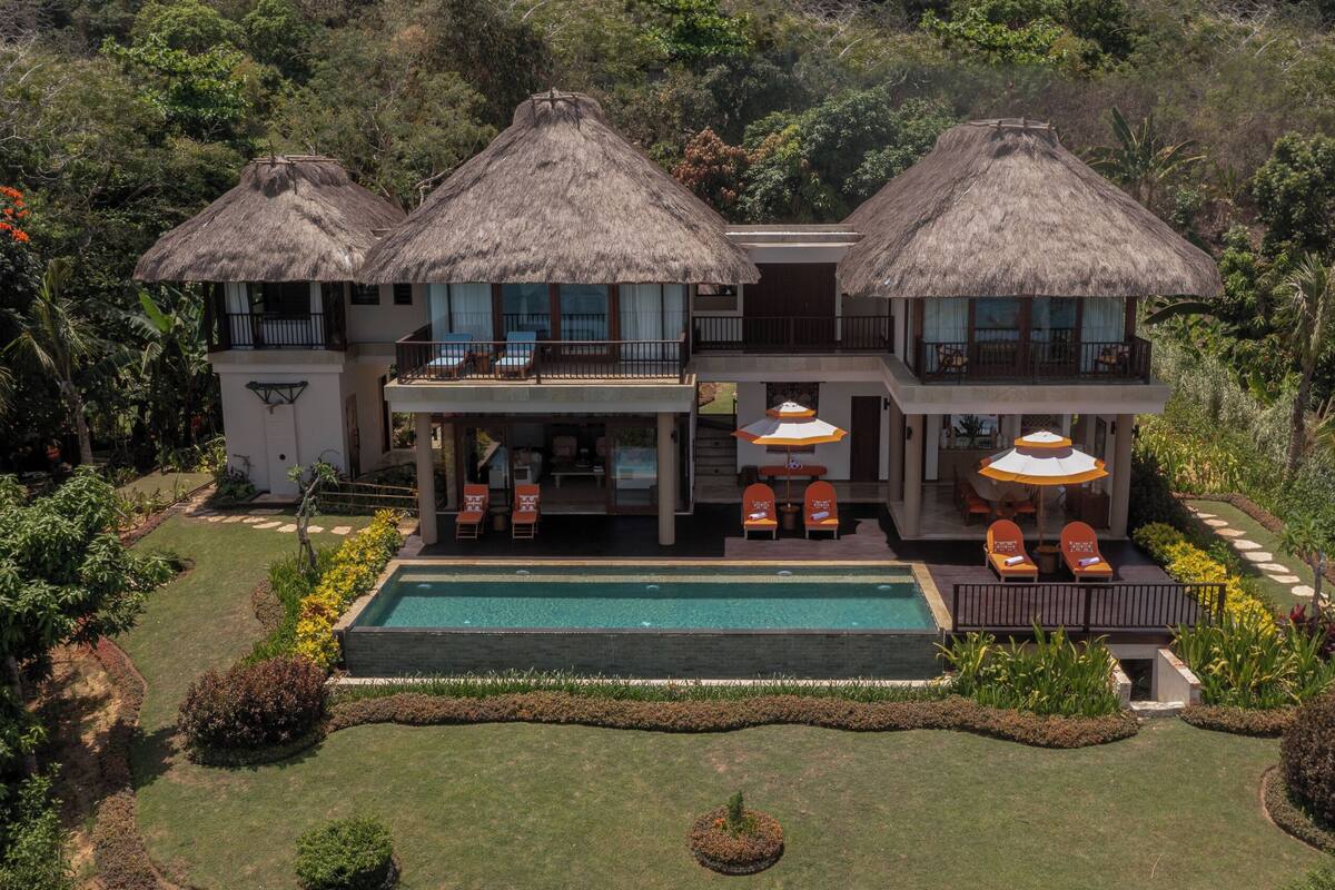 Une maison à deux étages avec une piscine, entourée d’une végétation luxuriante et de constructions aux toits de chaume.