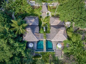 Vista aérea de un resort con villas de techo de paja, una piscina central y exuberante vegetación tropical.