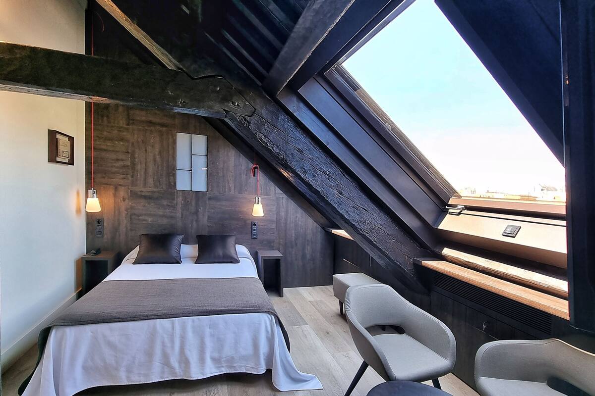 Un dormitorio con cama, dos mesitas de noche, una mesita auxiliar, escritorio, dos sillas, ventana con vistas y techo de madera.