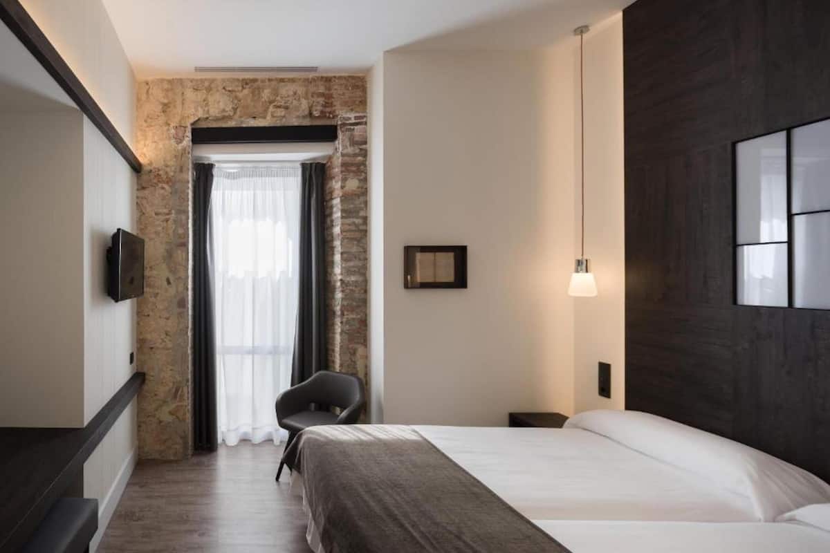 Habitación de hotel moderna con pared de acento de piedra, una cama grande, una silla y un televisor montado en la pared.