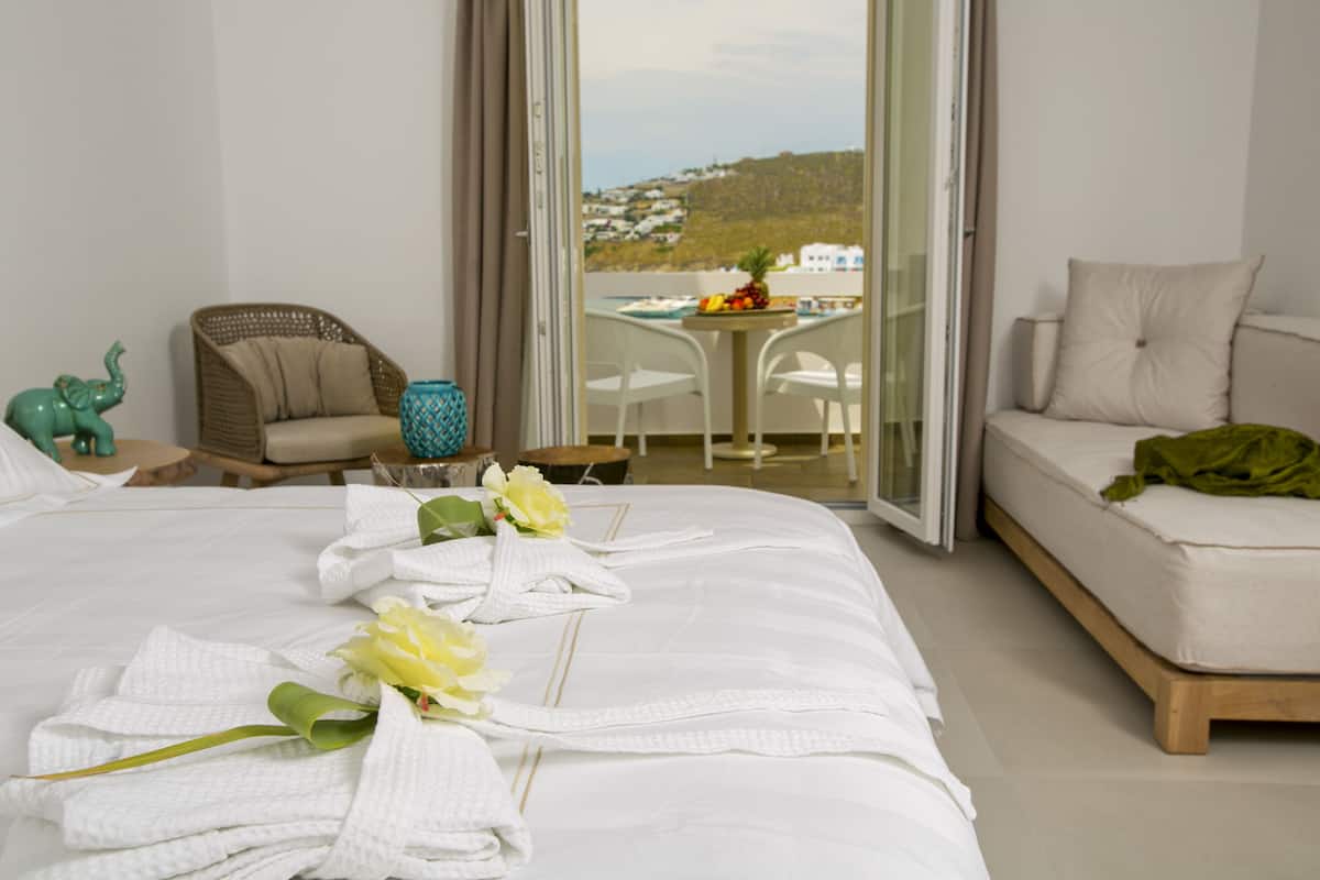 Ein Hotelzimmer mit Bett, einer Couch, einem Korbstuhl, einem Tisch mit Früchten und Blick auf eine Hügelkette.