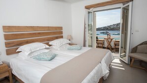 Un dormitorio con cabecera de madera, una cama grande con ropa de cama blanca, una manta beige y vista al mar a través de una puerta abierta.