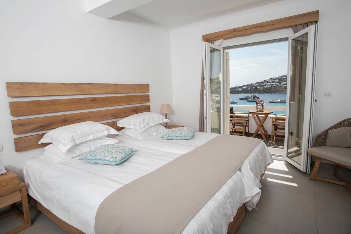 Ein Schlafzimmer mit einem hölzernen Kopfteil, einem großen Bett mit weißer Bettwäsche, einer beigen Tagesdecke und Meerblick durch eine offene Tür.