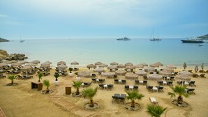 Una playa con filas de sillas de playa y sombrillas de paja, una vista despejada del mar y varios barcos fondeados cerca.