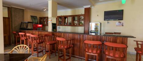 Zona de bar con taburetes de madera y una barra. Hay taburetes con asientos acolchados, una barra y un televisor montado en la pared.