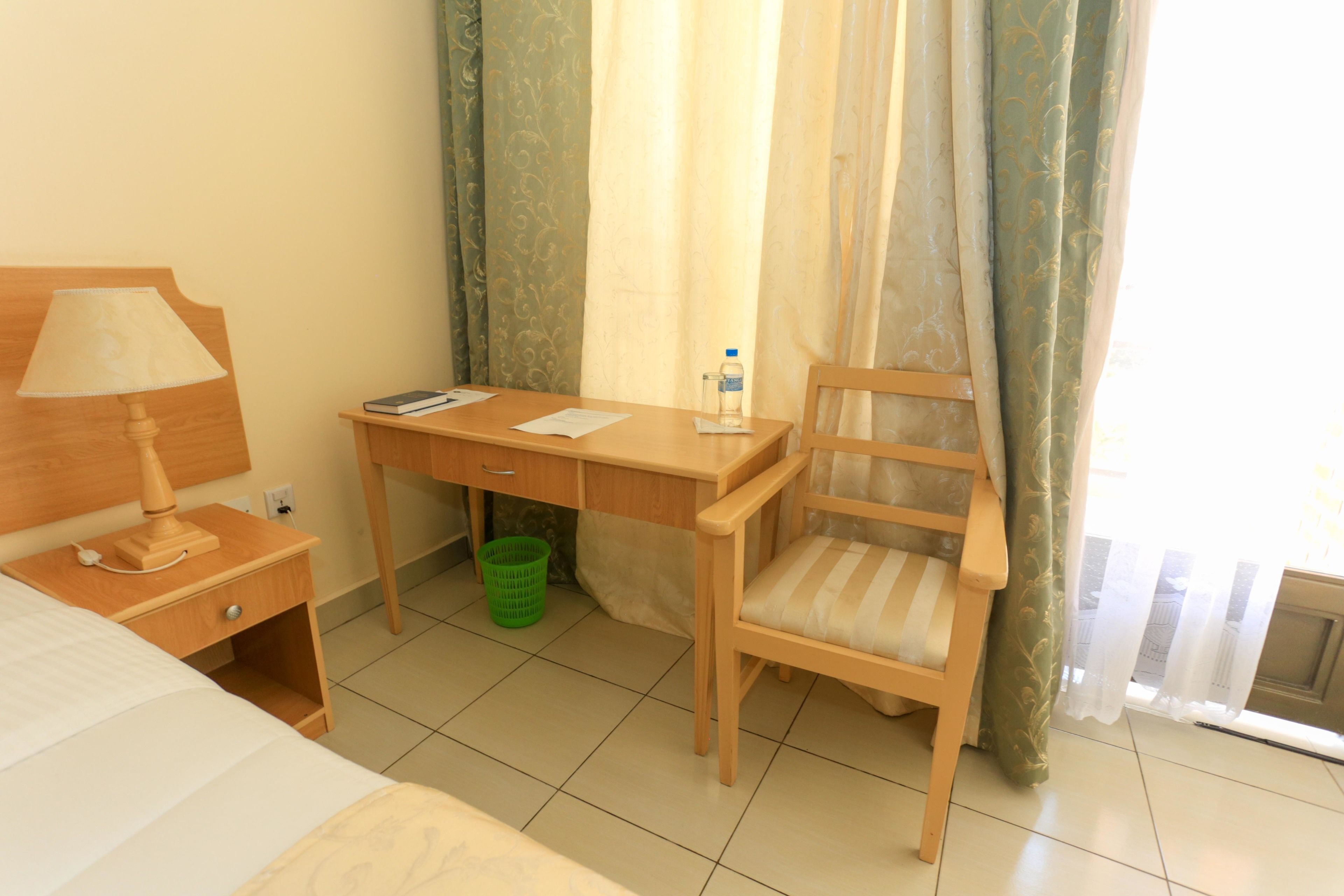 Habitación de hotel con cama, escritorio con lámpara, silla, basurero verde y ventana con cortinas.