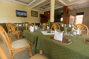 A dining area with tables set for four, each with a green tablecloth, white napkins, and cutlery. There are wooden chairs with cushioned seats. A television is mounted on the wall, and a painting is visible above the tables. - Faraja Hotel (Muhoza)