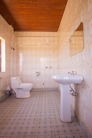 Un baño con papel tapiz estampado, un inodoro blanco, un lavabo sobre pedestal y techo de madera.