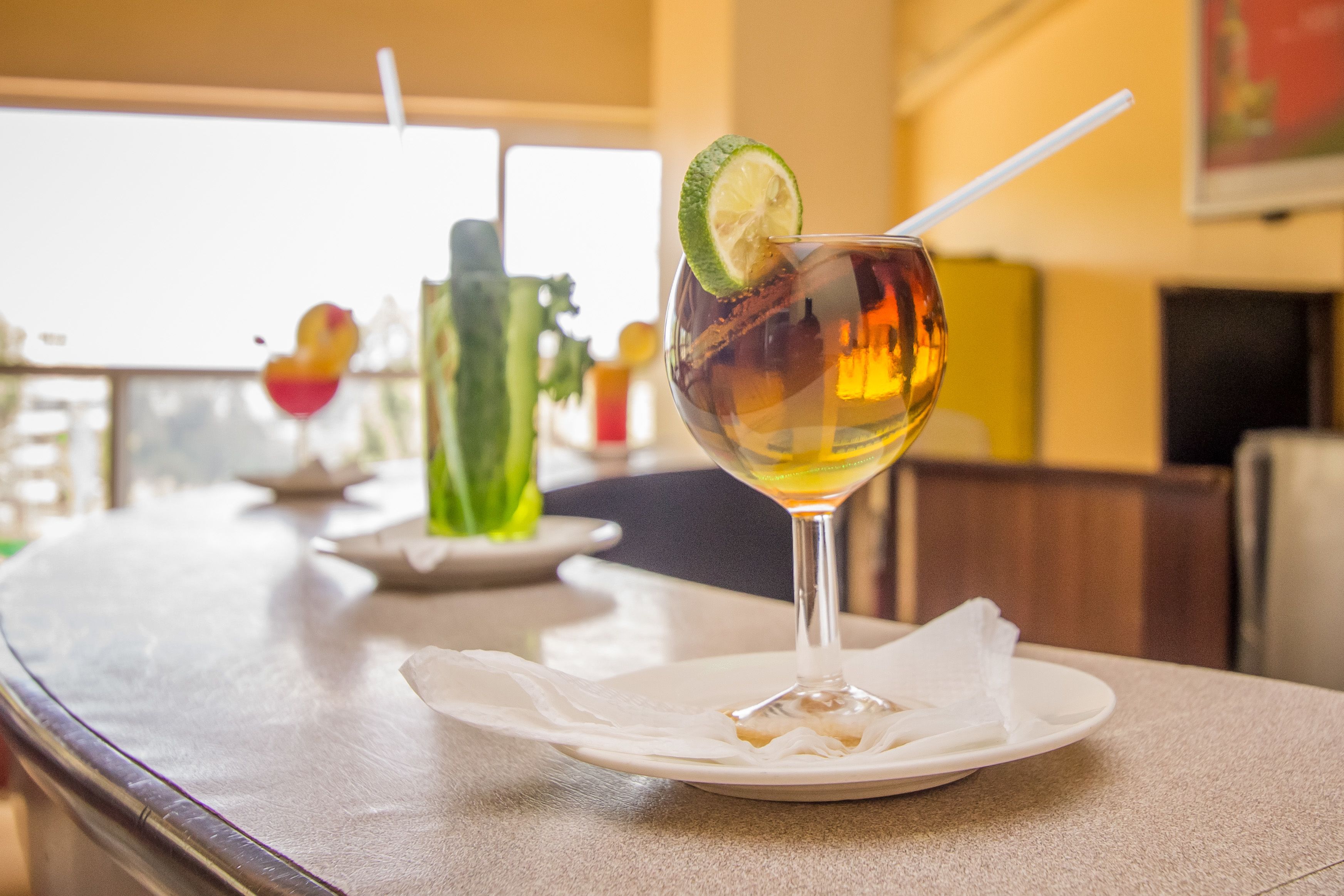 A table with a glass of amber-colored beverage garnished with a lime slice and a straw, a white napkin, and a plate with a small item.