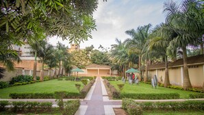 Uma passagem leva a um edifício com área coberta, cercado por vegetação bem cuidada e palmeiras.