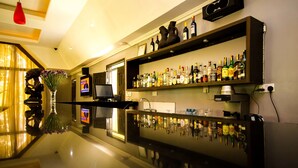 Zona de bar con mostrador pulido, estanterías llenas de botellas de licor, un televisor y un jarrón con flores.