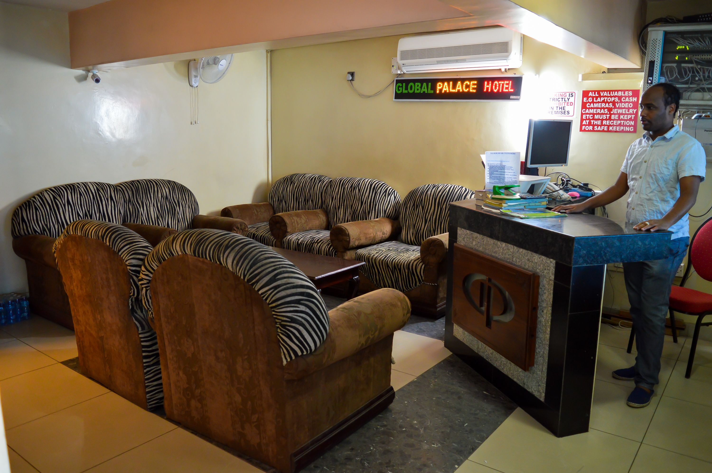 A hotel reception area with a counter, several zebra-striped sofas, a wall-mounted air conditioner, and a sign indicating the hotel name.