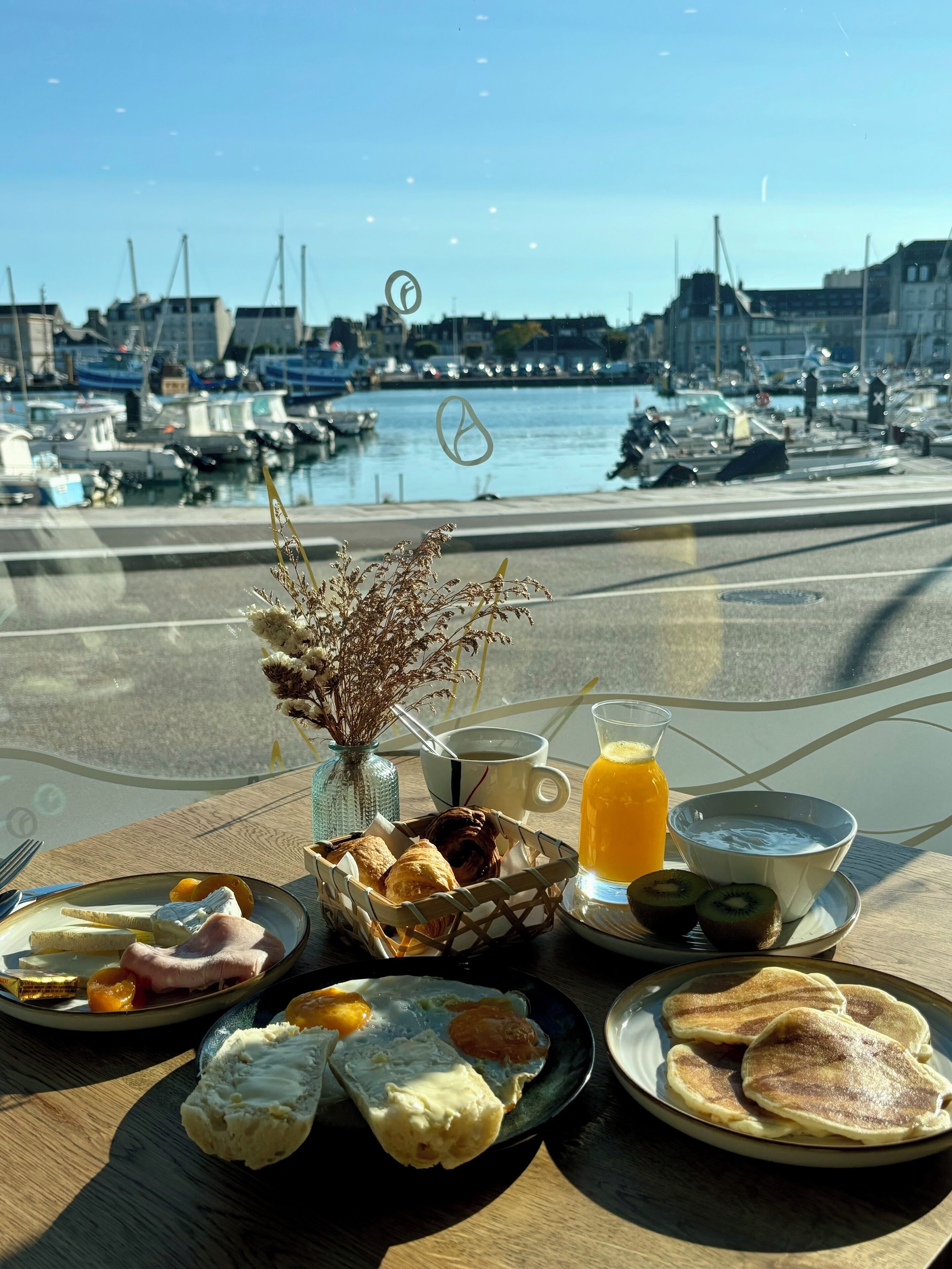 A breakfast table with various foods and a glass of orange juice, overlooking a marina with boats and buildings.