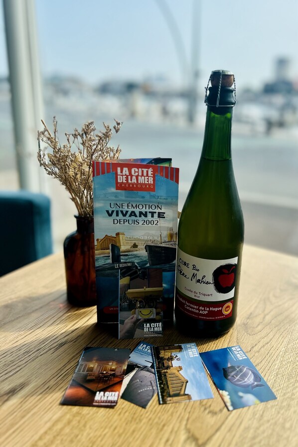 A bottle of wine, a book titled 'LA CITÉ DE LA MER', and brochures on a wooden table with a view of the sea in the background. - Hôtel Ambassadeur (Cherbourg-en-Cotentin)