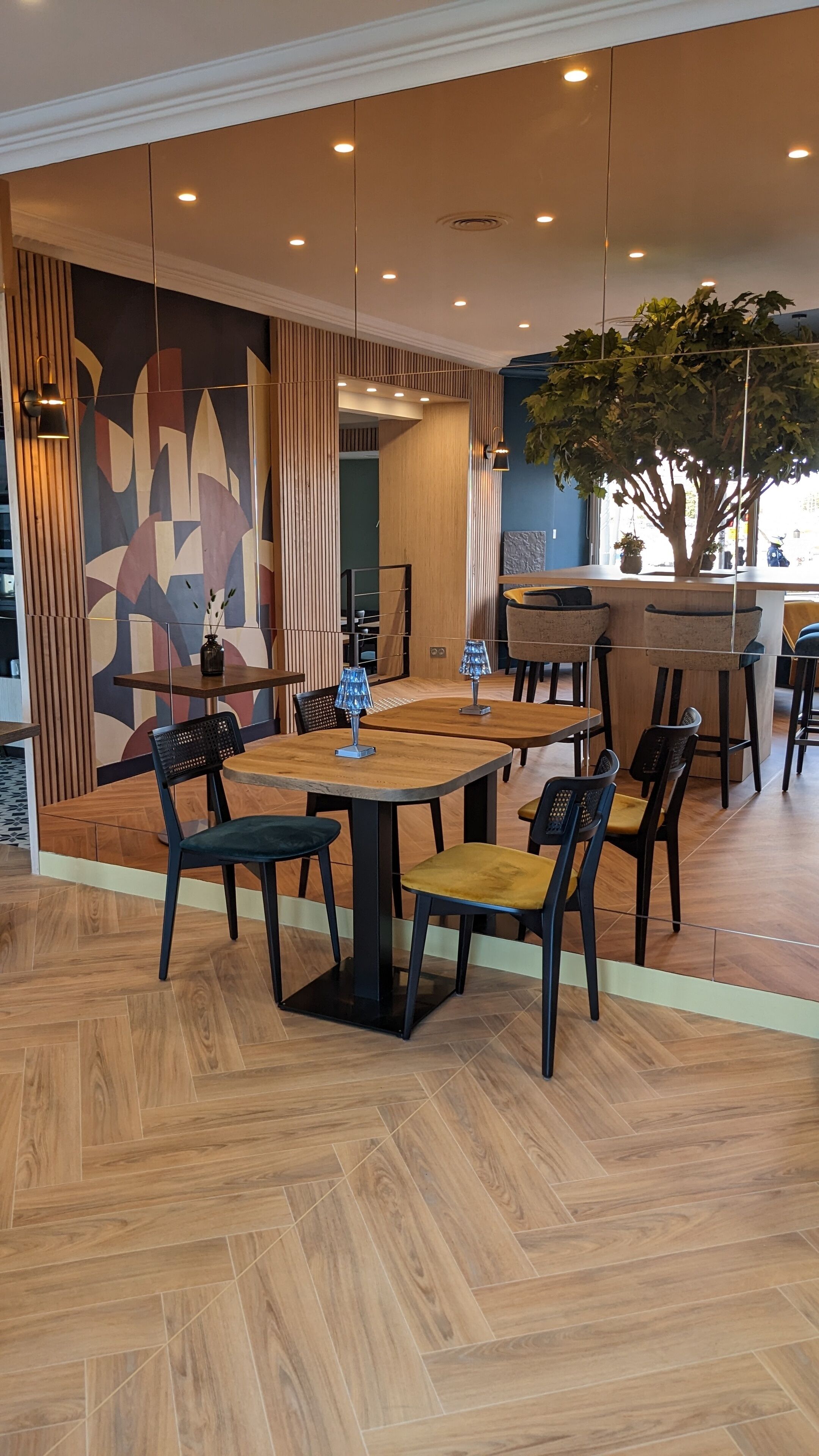 Un comedor moderno con mesas y sillas de madera, una pared decorativa con patrones geométricos y una planta grande en maceta.