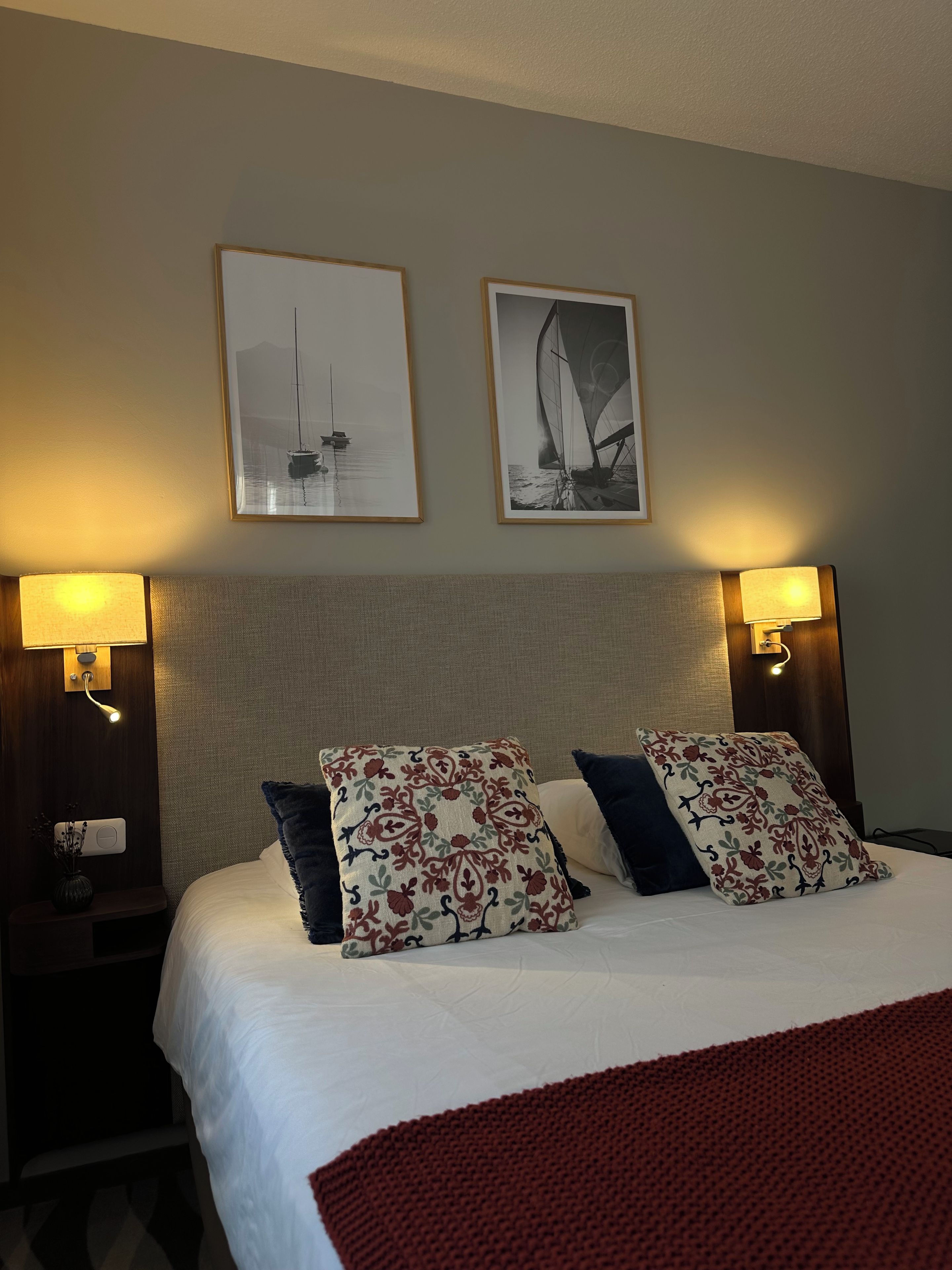 Una habitación de hotel con una cama, dos cuadros enmarcados, dos lámparas de noche y una mesita de noche con un florero.