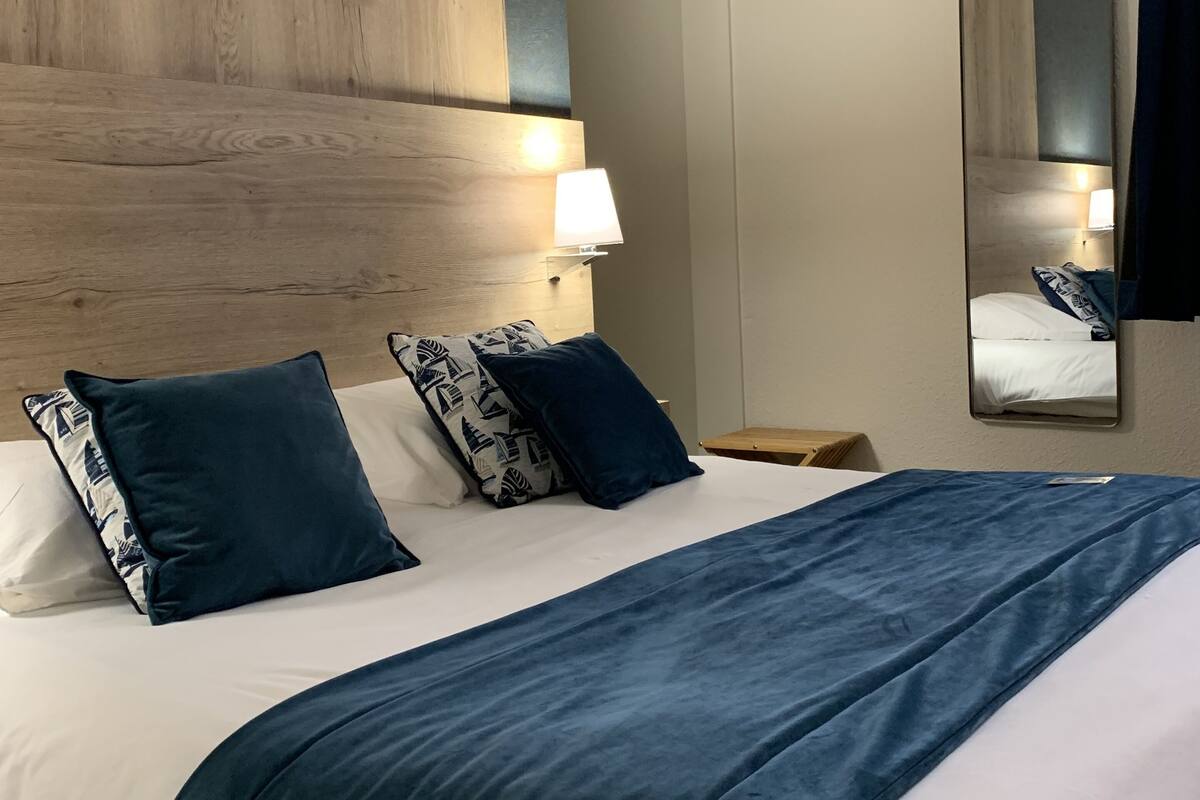 A hotel room with a wooden headboard, a large bed with a blue bedspread, multiple pillows, a bedside lamp, and a mirror on the wall.