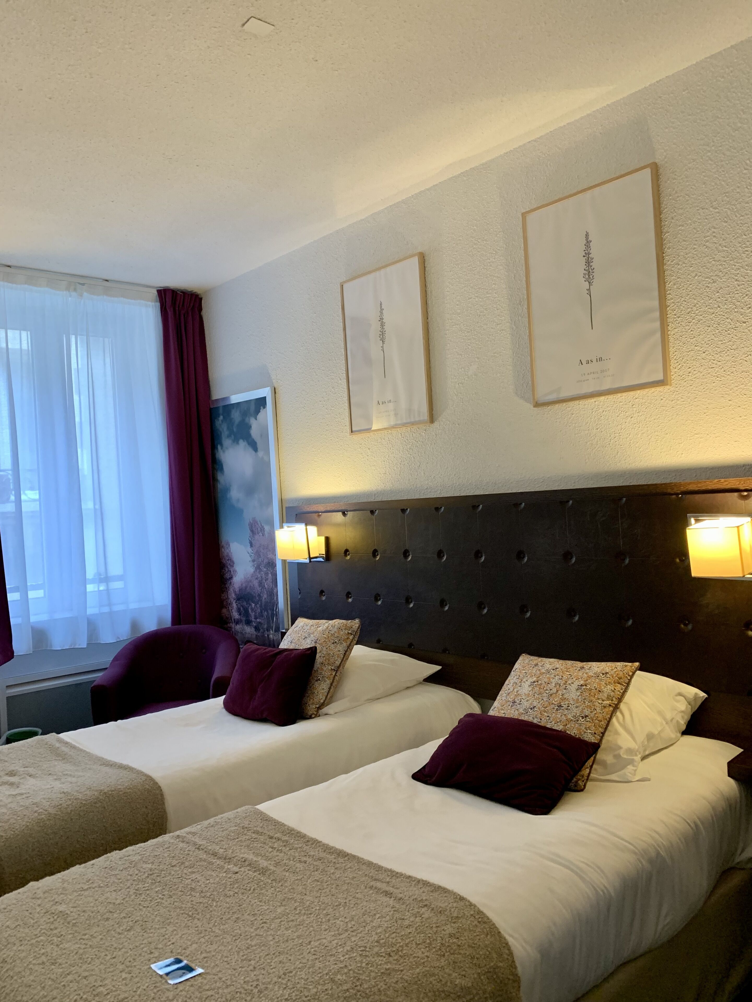 A hotel room with two beds, a large window with sheer curtains, a dark headboard, and framed artwork on the wall.