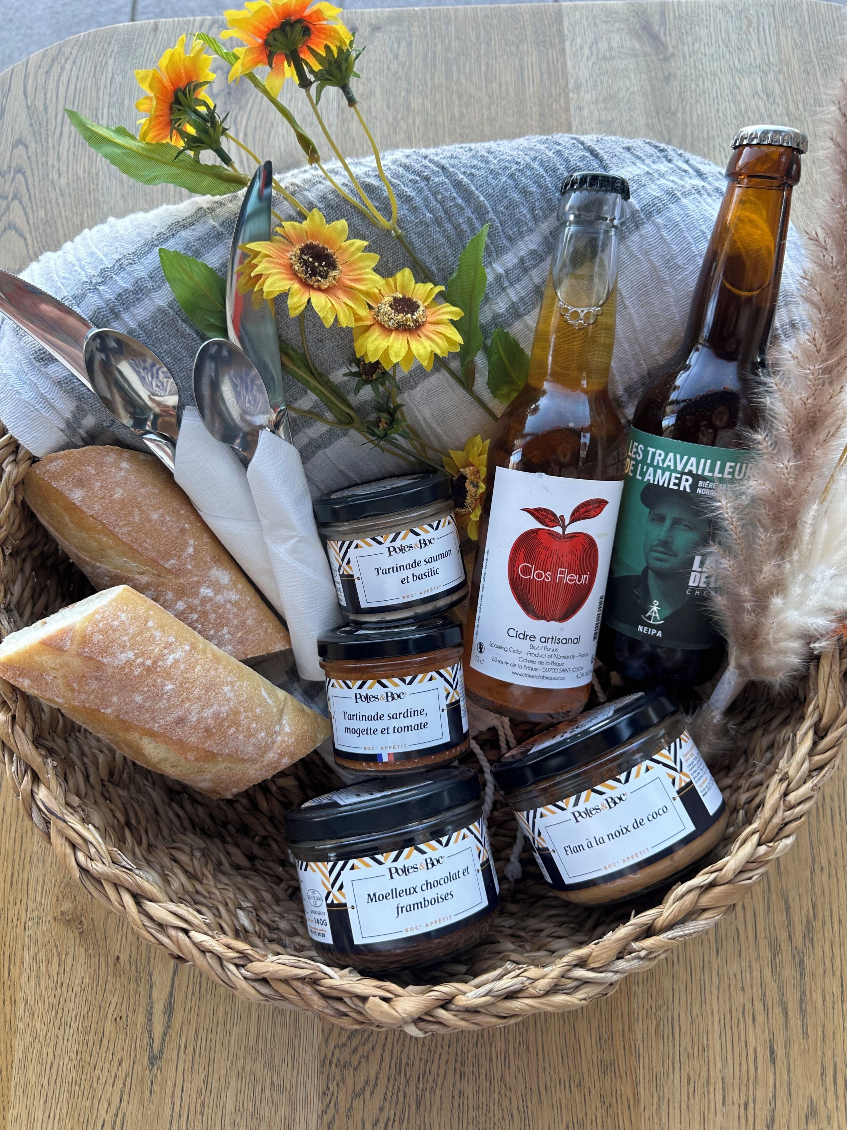 Una cesta de mimbre con frascos de alimentos en conserva, una hogaza de pan, una botella de cerveza, un par de tijeras y un jarrón con girasoles.